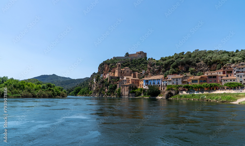Idyllic riverside village with terracotta-roofed houses nestled on a lush hillside, crowned by a majestic fortress, under the azure sky. Idyllic riverside village with terracotta-roofed houses nestled on a lush hillside, crowned by a majestic fortress, under the azure sky.
