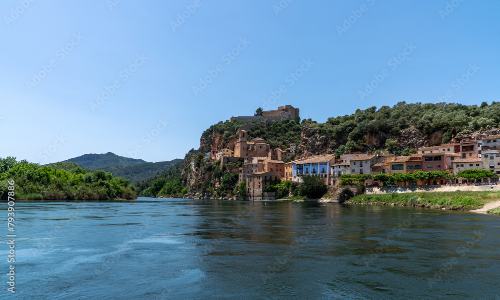 Idyllic riverside village with terracotta-roofed houses nestled on a lush hillside, crowned by a majestic fortress, under the azure sky.