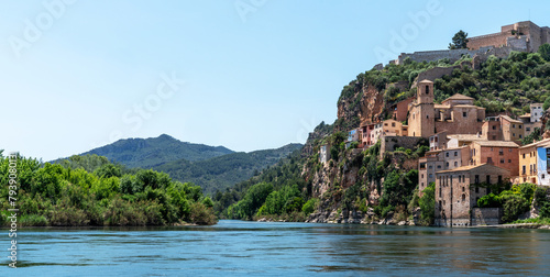 a picturesque riverside village Miravet, Spain. Ancient buildings clinging to the steep hillside, the river flows calmly in the foreground, framed by the lush greenery of the surrounding landscape.. photo