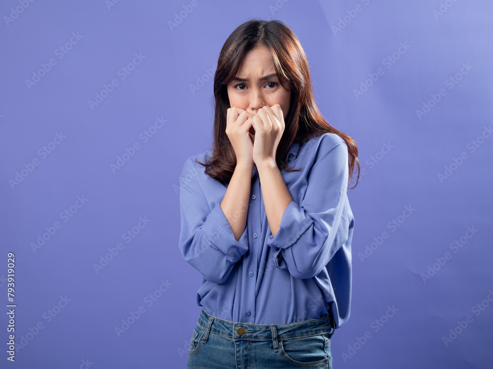 A young Asian woman in a light blue button-up shirt and jeans stands against a solid lavender background, nervously biting her fingernails with a worried expression, conveying anxiety or fear. A young Asian woman in a light blue button-up shirt and jeans stands against a solid lavender background, nervously biting her fingernails with a worried expression, conveying anxiety or fear.