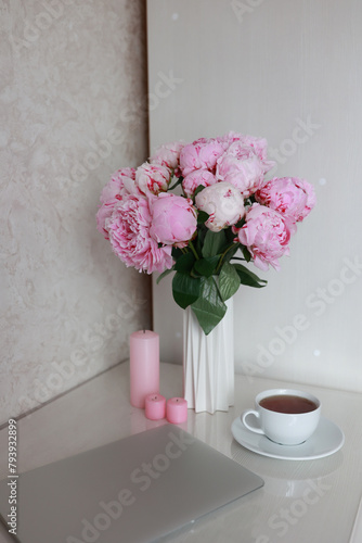 tea in a white cup, pink peonies, flowers and candles of pink color on a white background.Laptop, beautiful pink peony bouquet and notebook on white table background. minimalist home workspace. 