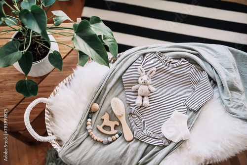 Still life background of cute baby products - changing basket with baby bodysuit, newborn clothes, knitted rabbit and wooden toy. Minimalist style photography of baby shower, pregnancy announcement.