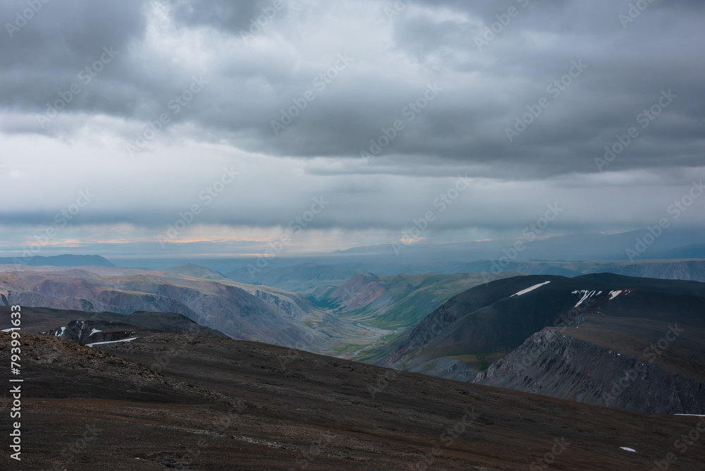 Dramatic top view from precipice edge to wide colorful valley and mountain silhouettes in rain under lead gray cloudy sky. Dark atmospheric mountains in rainy weather and sunset landscape far away.