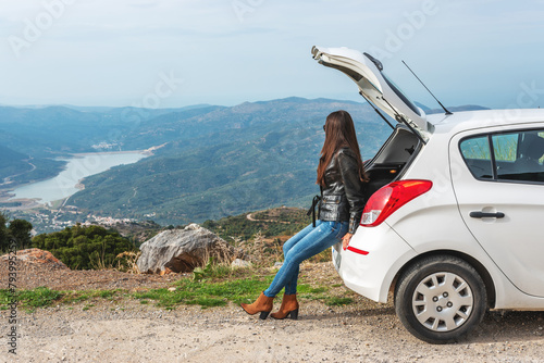 Woman traveler sitting on hatchback car with mountain background.