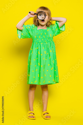 Investigator researcher scientist school girl holding magnifying glass near face looking into camera with big zoomed funny eye searching, analysing. Preteen child kid on yellow background. Vertical