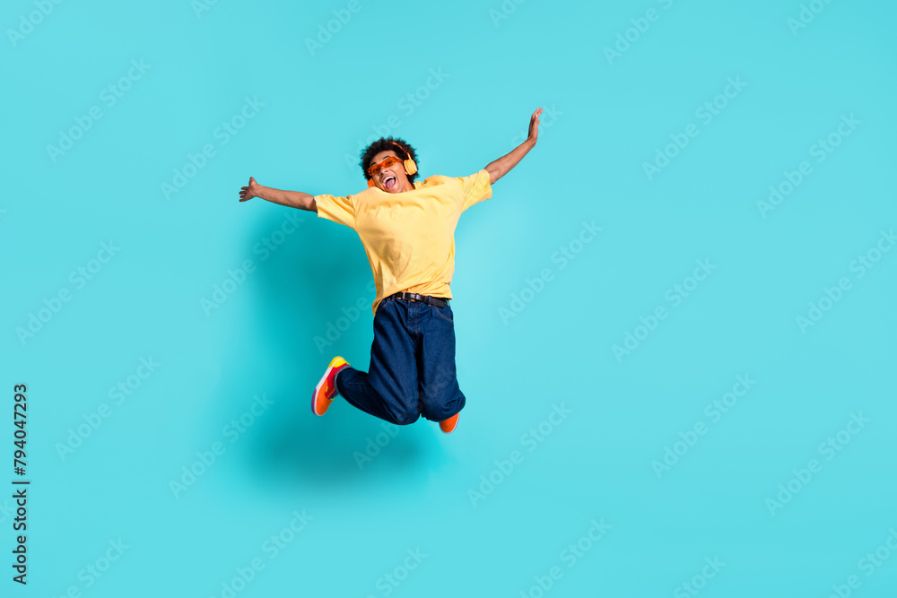 Full length photo of ecstatic guy dressed yellow t-shirt jeans jumping in headphones hands up isolated on turquoise color background
