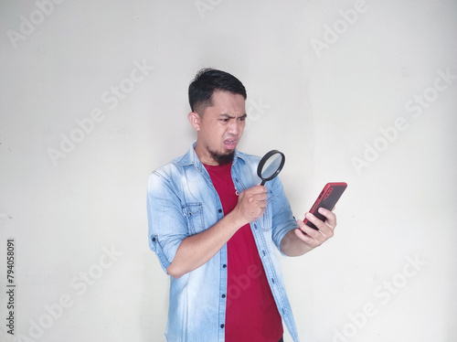 Adult Asian man looking to mobile phone using magnifying glass with suspicious expression photo