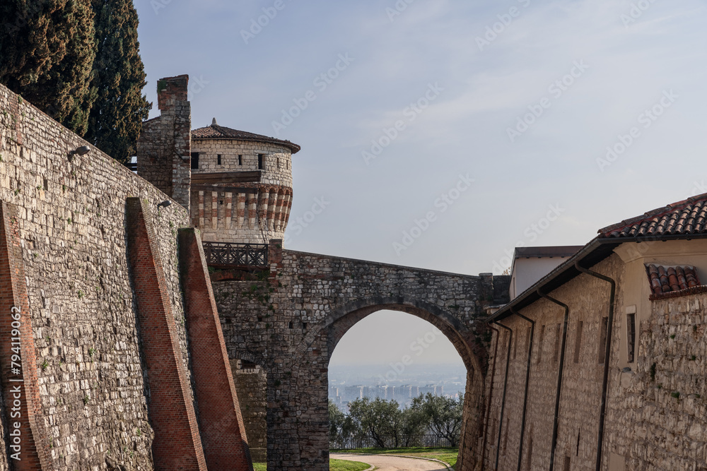 View from Brescia historic castle grounds, where the aged stone bridge arch leads to the Torre dei Prigionieri, overlooking the distant cityscape