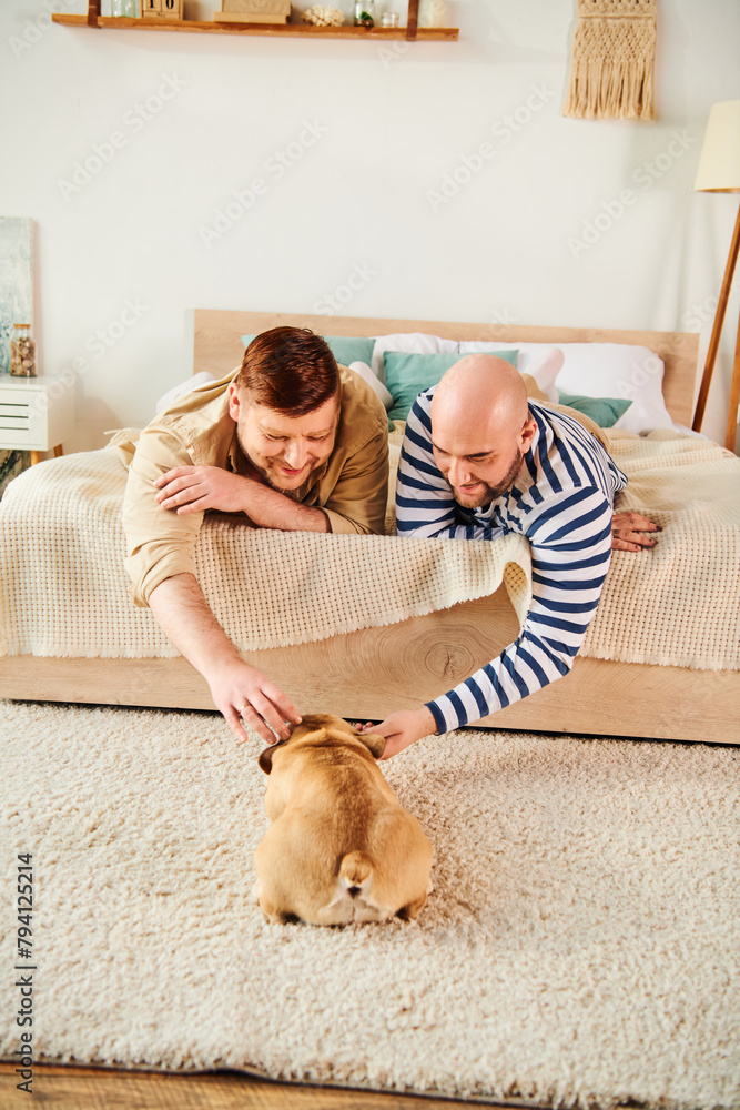Two men laying on a bed next to a French bulldog, relaxing together.