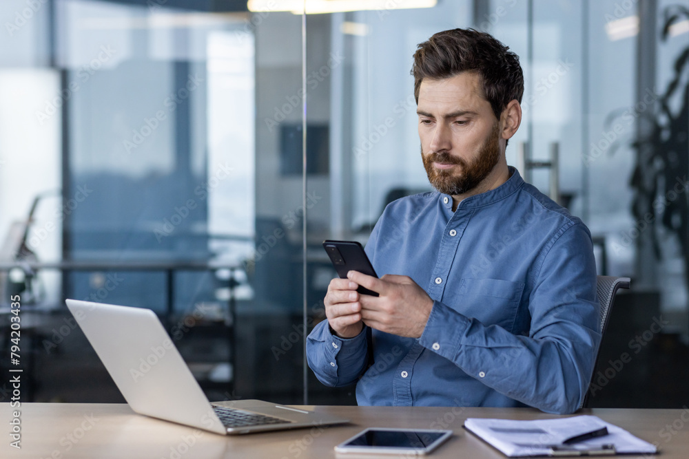 A serious young man sits in the office at the desk and uses a mobile phone in his hands