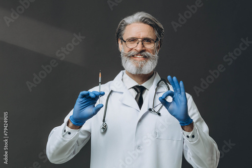 Senior doctor with grey hair holding syringe doing ''Ok'' sign with fingers, smiling friendly.
