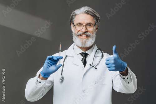 A senior doctor with gray hair is holding a syringe and giving a thumbs up, smiling warmly.