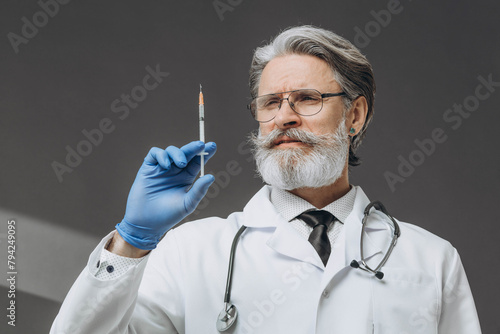 Portrait of a senior doctor in gloves and medical gown holding a syringe. isolated on gray background.