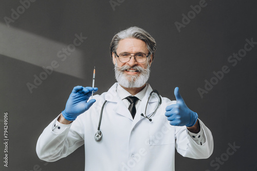 A senior doctor with gray hair is holding a syringe and giving a thumbs up, smiling warmly.