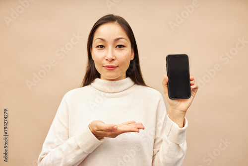 Cute happy smiling asian girl presenting new gadget or mobile application holding smartphone with black copy space for your advertising content and showing at it with hand, isolated on pink background