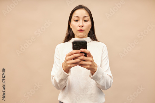 Studio image of focused concentrated beautiful asian girl holding smartphone and reading news, chatting online, replying on message in corporate chat, searching for information, surfing internet