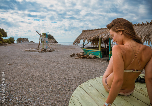 woman in bikini sitting on wooden pier by the beach with rustic huts and cloudy sky"