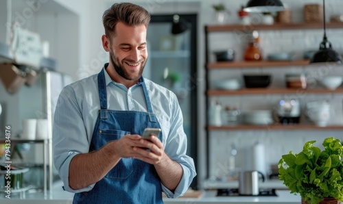 A handsome smiling man holding a cell phone in his hand standing in his kitchen photo