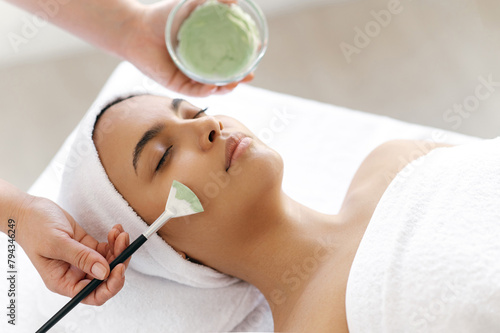 Skincare, beauty procedure. Therapist applying green face mask on the face of a beautiful brazilian or hispanic young woman. Facial treatments, wrinkle prevention, facial cleansing