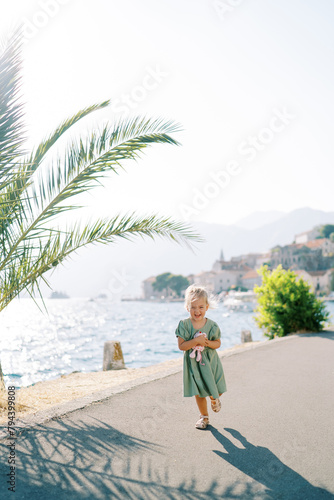 Little laughing girl with a toy walks along the embankment past a palm tree