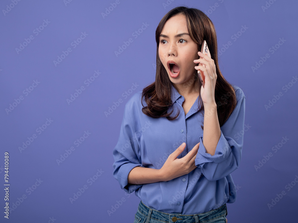 Shocked Asian woman in blue blouse reacting during a phone call, expressive and surprised, against a plain purple background Shocked Asian woman in blue blouse reacting during a phone call, expressive and surprised, against a plain purple background