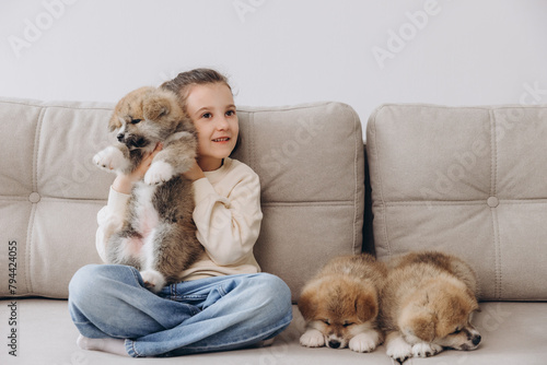 Little happy smiling girl playing and hugging Akita Inu puppies at home on sofa