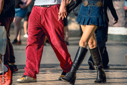 Urban stride, a young man and a girl, hand in hand, walk in sync along a busy street, their lower halves in focus. Red trousers meet denim skirt, a snapshot of city life and fashion