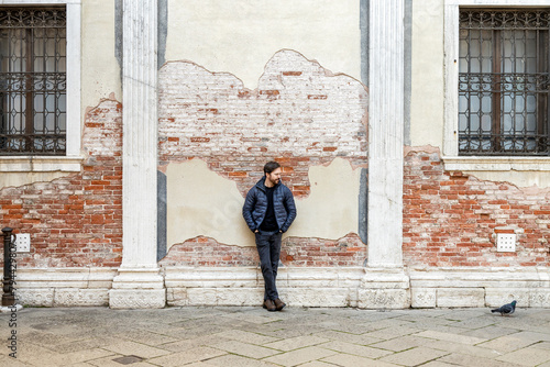 An elegant bearded man wearing casual clothes stands leaning against wall with hands in his pockets looking at a pigeon in Venice, Italy. Old venetian grungy brick wall with damaged plaster