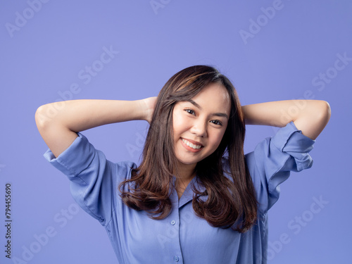 Portrait of an Indonesian Asian woman, wearing a blue shirt, smiling with her hands behind her head, isolated against a purple background