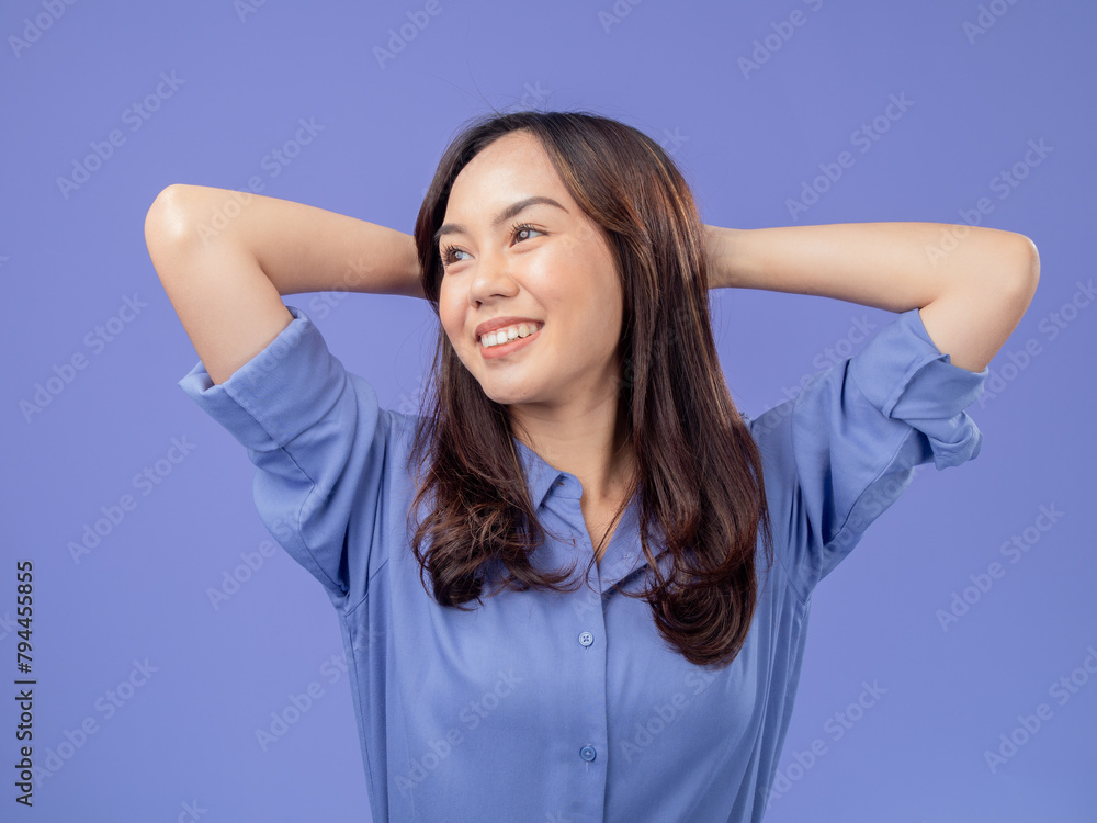 Portrait of an Indonesian Asian woman, wearing a blue shirt, smiling with her hands behind her head, isolated against a purple background Portrait of an Indonesian Asian woman, wearing a blue shirt, smiling with her hands behind her head, isolated against a purple background