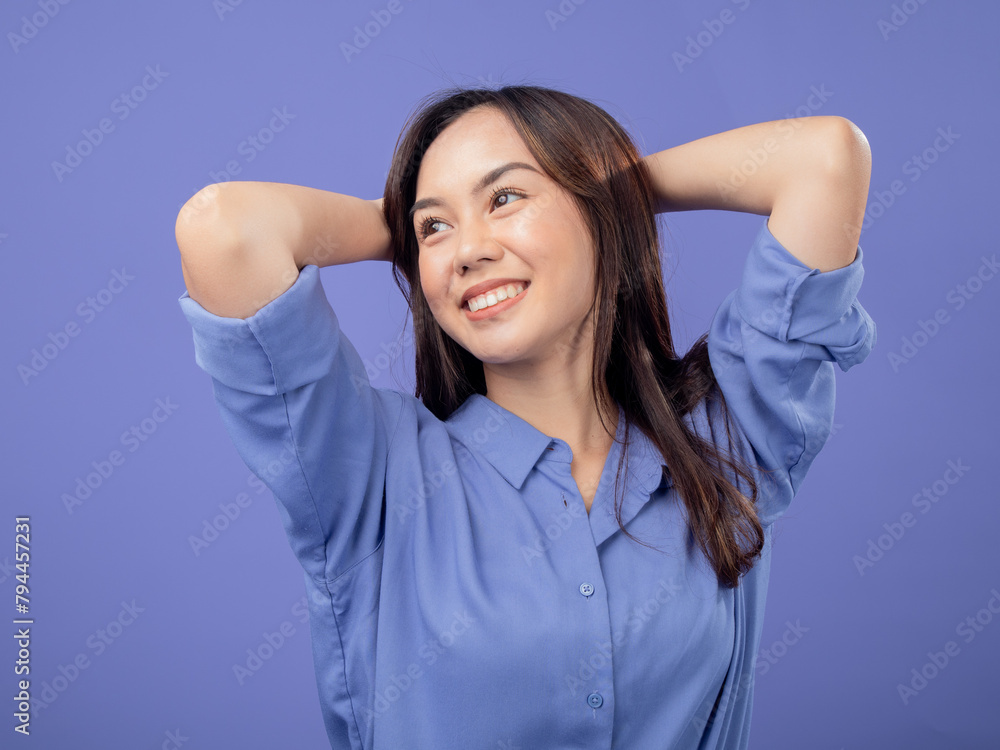 Portrait of an Indonesian Asian woman, wearing a blue shirt, smiling with her hands behind her head, isolated against a purple background Portrait of an Indonesian Asian woman, wearing a blue shirt, smiling with her hands behind her head, isolated against a purple background