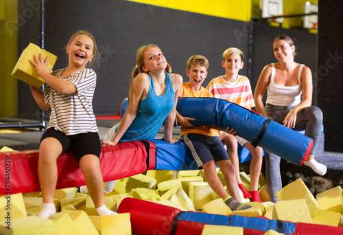 Group of happy tween children and young woman sitting on soft beam above pit of foam cubes, relaxing while playing together in indoor amusement park