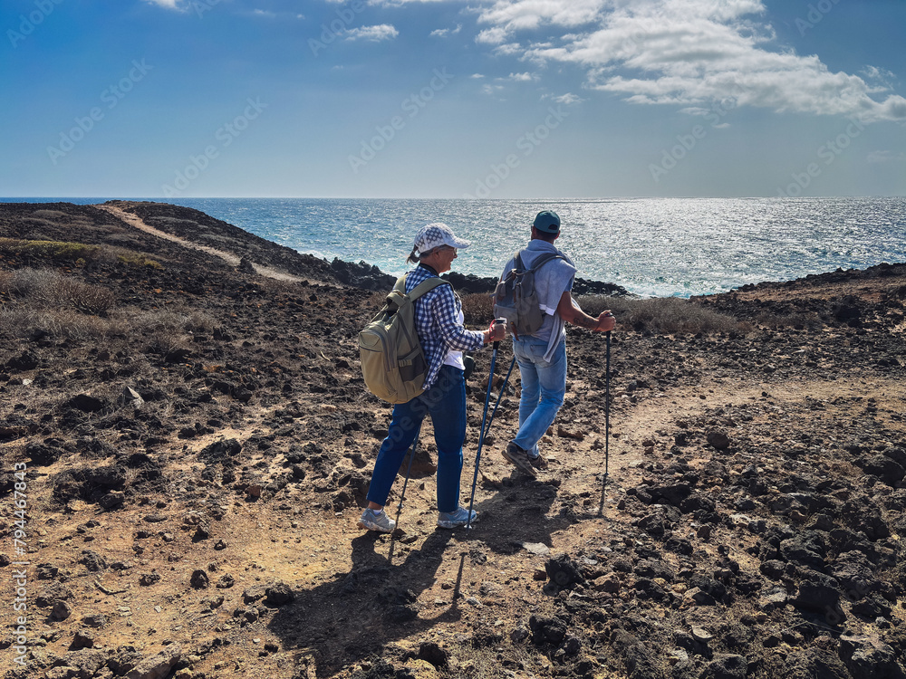Outdoor activity in nature concept. Senior woman and middle aged son walking together in arid footpath along the sea enjoying healthy lifestyle and freedom. Blue sky and horizon over water