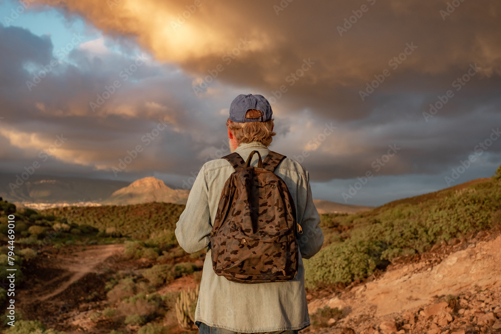 Back view of senior man with hat and casual clothing walking outdoors in sunset light with dramatic sky. Healthy lifestyle, vacation and sporting activity in retirement Back view of senior man with hat and casual clothing walking outdoors in sunset light with dramatic sky. Healthy lifestyle, vacation and sporting activity in retirement
