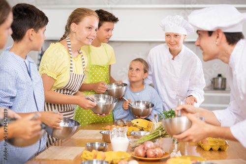 Friendly enthusiastic tween girl learning how to cook and listening to advices of professional chef during group cooking masterclass ..