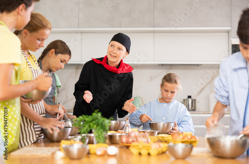 Friendly professional female chef in black uniform giving culinary lesson to group of interested tweens, teaching cooking skills..