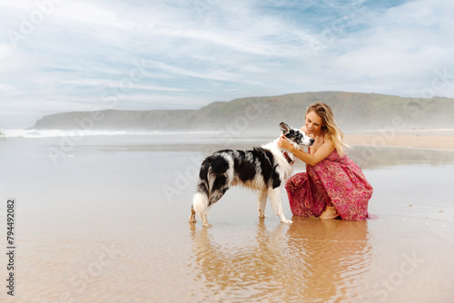 blonde girl crouching in the sand petting and cuddling her border collie dog during the summer.