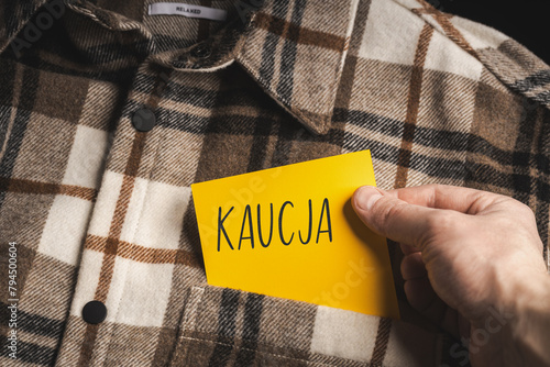 Yellow card with a handwritten inscription "Kaucja", held in the hand against the background of a brown plaid shirt (selective focus), translation: deposit