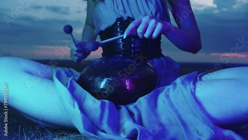Young performer plays the hapi drum in blue neon light, against the backdrop of an inspiring and meditative evening sky. She are sitting on the grass, in the lotus position, surrounded by nature photo