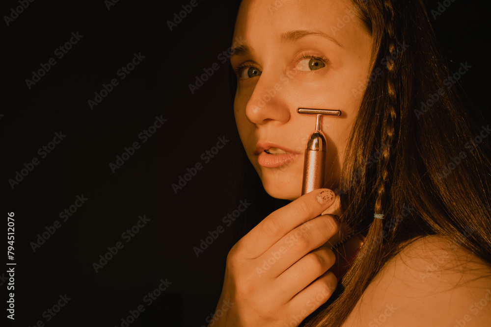 Obraz premium Young beautiful white woman, girl is using vibrating golden facial massager on black background. Face massage. Natural beauty youth Female skin care.