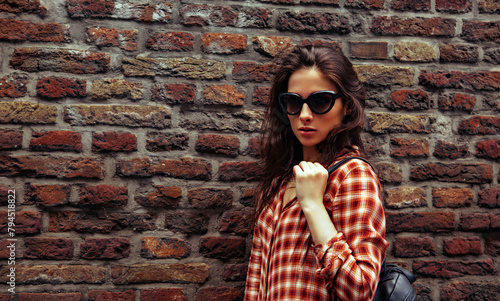 Beautiful brunette woman with long brown hair in fashion sunglasses standing on the old red brick wall building background in casual red shirt on the spring city. Closeup vintage banner