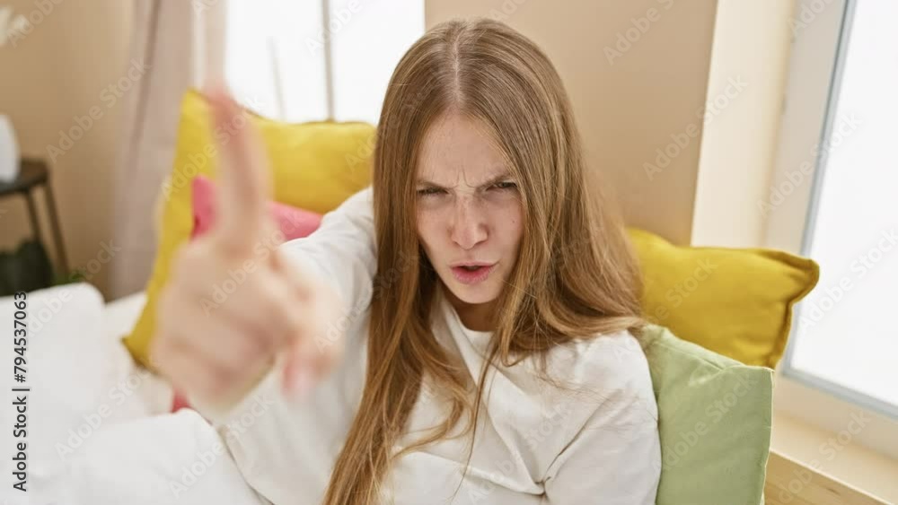 Angry young blonde woman in pyjama, furiously pointing at camera in displeasure, sitting on her bedroom bed, furious, unhappy and accusing you Angry young blonde woman in pyjama, furiously pointing at camera in displeasure, sitting on her bedroom bed, furious, unhappy and accusing you