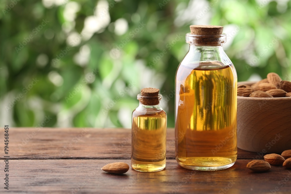Almond oil in bottles and nuts on wooden table against blurred green background, closeup. Space for text
