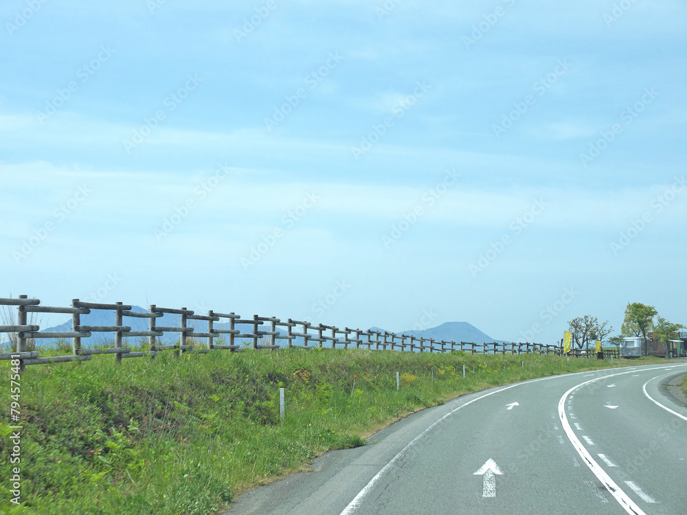 Travelling on Kyushu, driving to distant Mount Aso famous volcano caldera contour on lonely road with lush green grass valley and wooden fence on roadside