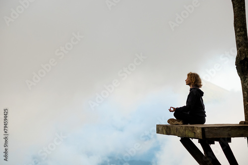 A young girl of European appearance meditates and does yoga on a wooden platform in the mountains, against the backdrop of clouds.