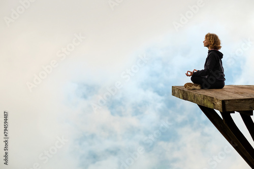 A young girl of European appearance meditates and does yoga on a wooden platform in the mountains, against the backdrop of clouds.