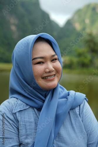 Asian model in hijab wearing blue shirt is posing in nature with mountains and water in the countryside as a backdrop