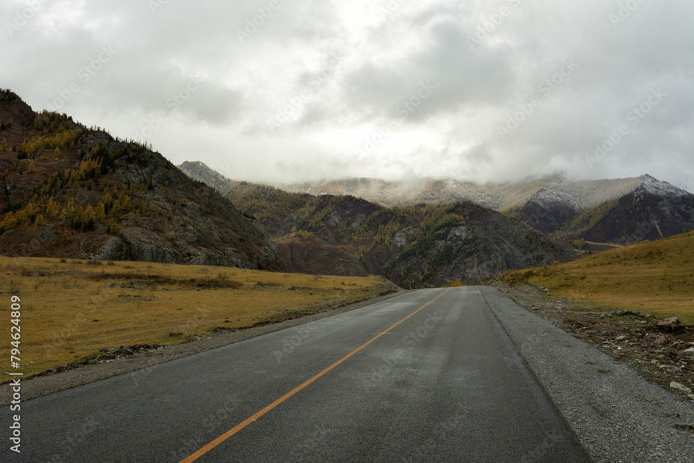 A straight asphalt road crosses a wide clearing between two high mountains on a cloudy autumn day.
