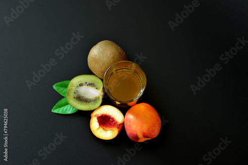 Peach juice with kiwi in a tall glass on a black background, next to pieces of ripe fruit with leaves.
