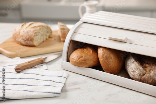 Wooden bread basket with freshly baked loaves and knife on white marble table in kitchen photo
